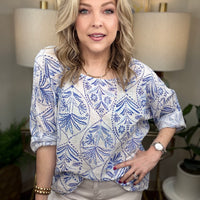 Woman wearing a blue and white patterned blouse in a home setting with lamps and plants.