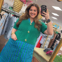 Woman taking a mirror selfie in a clothing store wearing a green top and blue striped skirt.