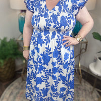 A woman with blonde hair wearing a white and blue floral print maxi dress with smocked bodice and ruffled cap sleeve
