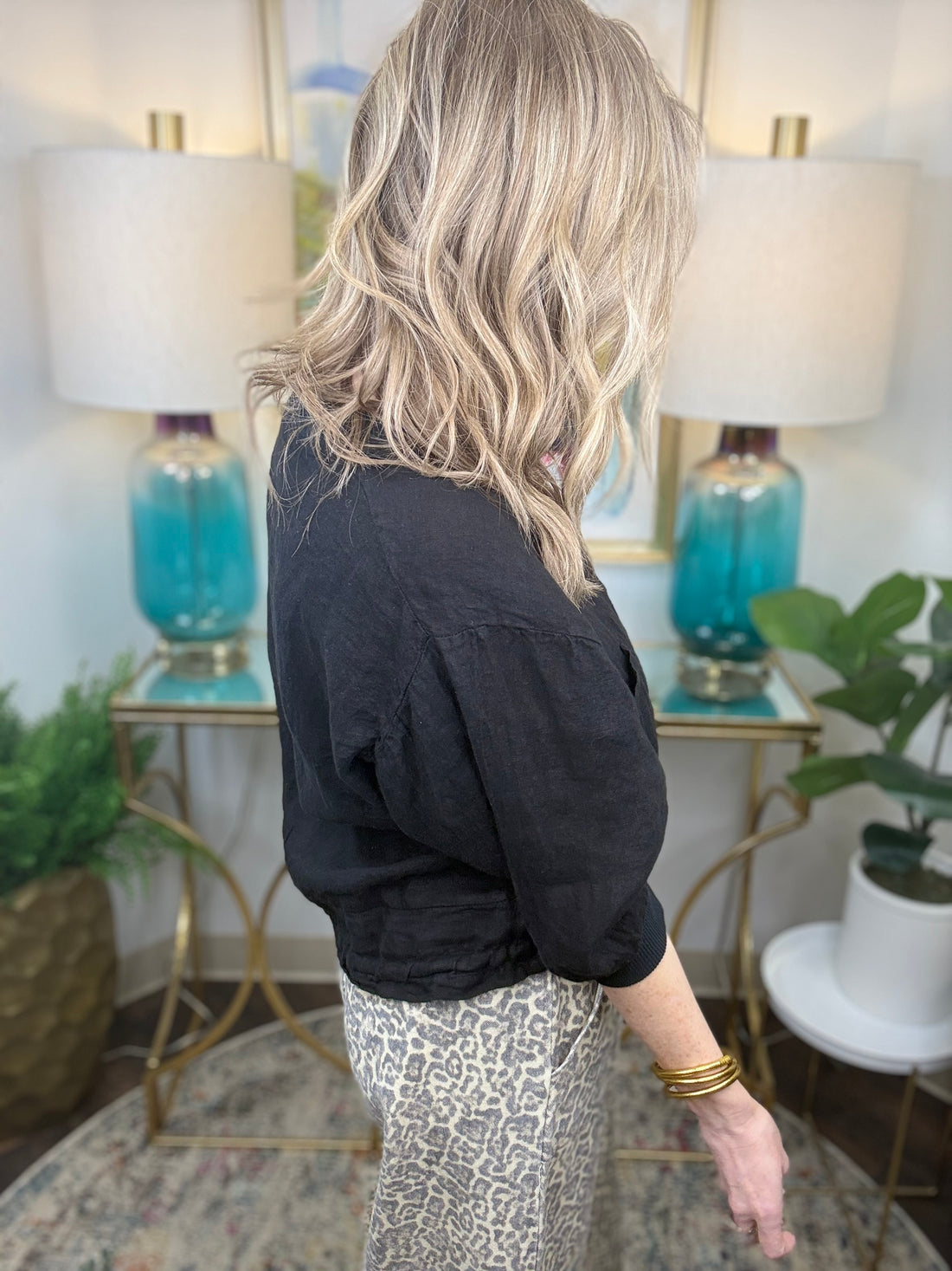 Woman in a black top and patterned pants standing in a room with lamps and plants.