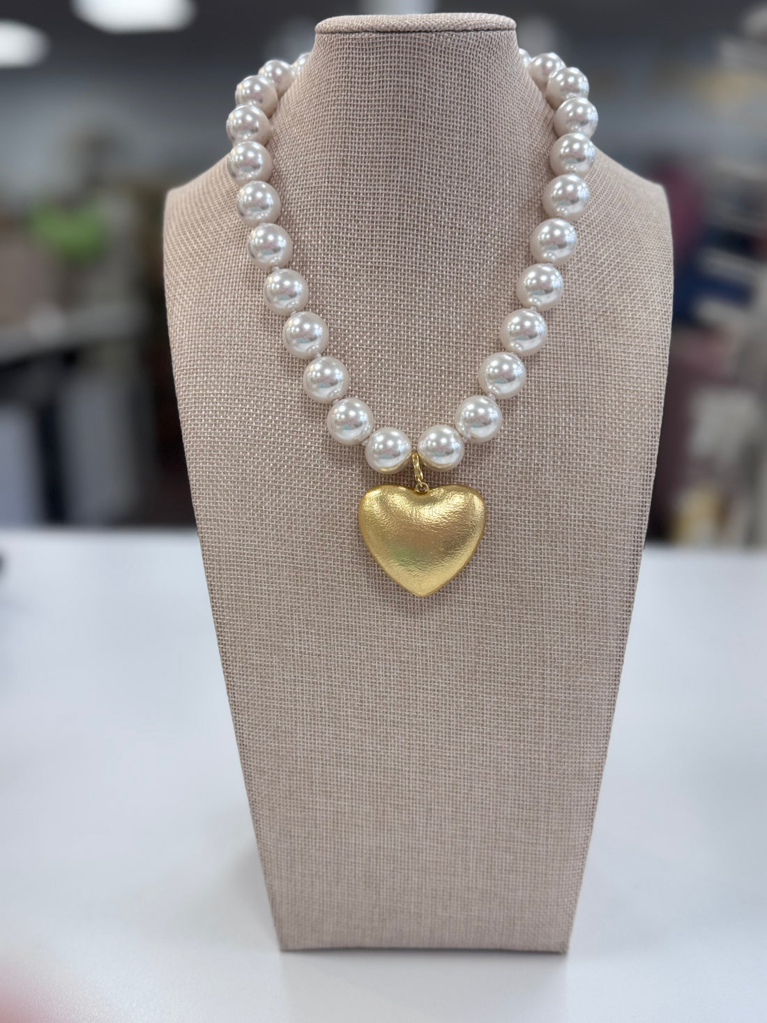 Pearl necklace with a gold heart pendant on a beige display stand.