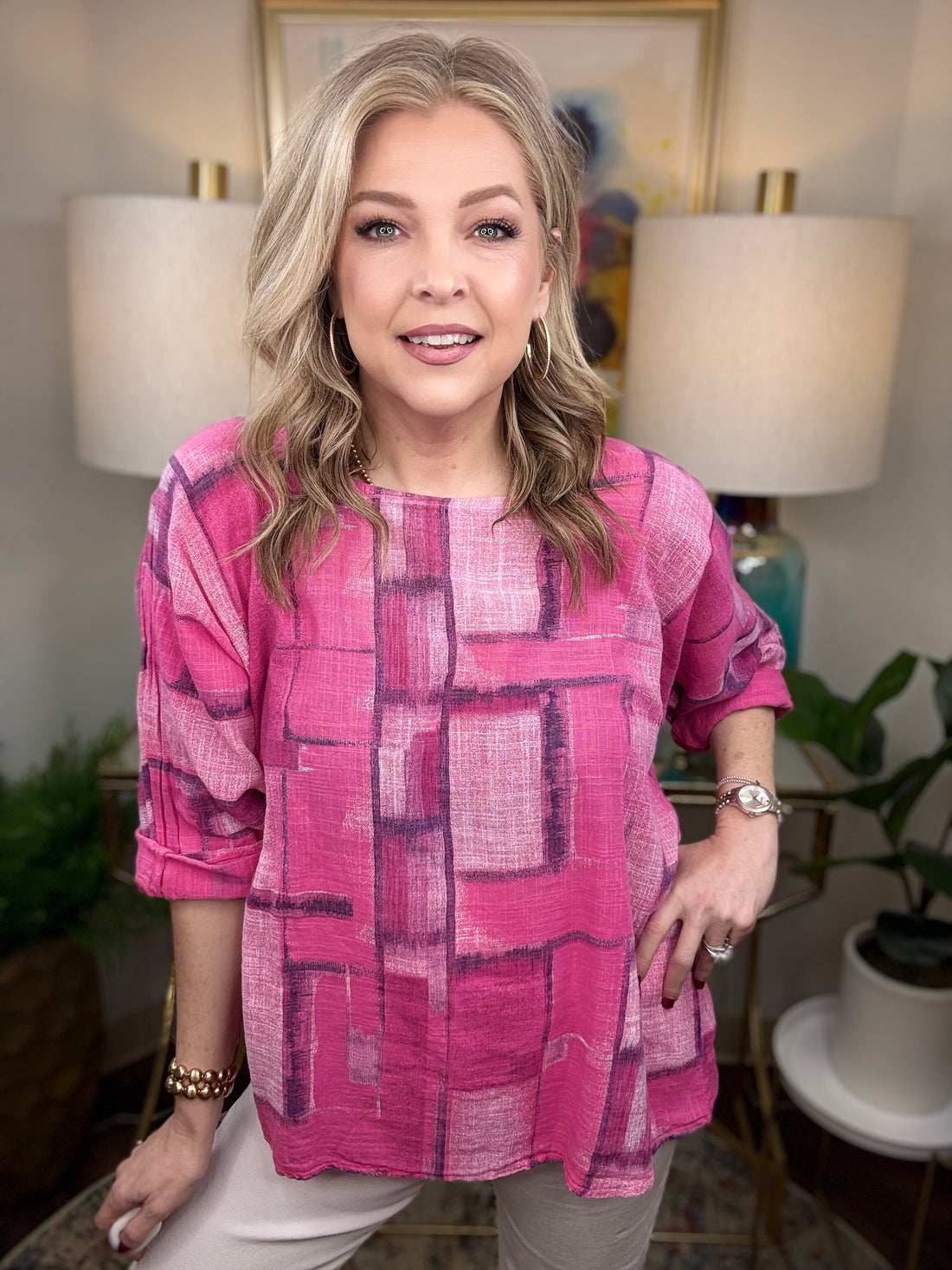 Woman wearing a pink patterned blouse in a home setting with lamps and plants.