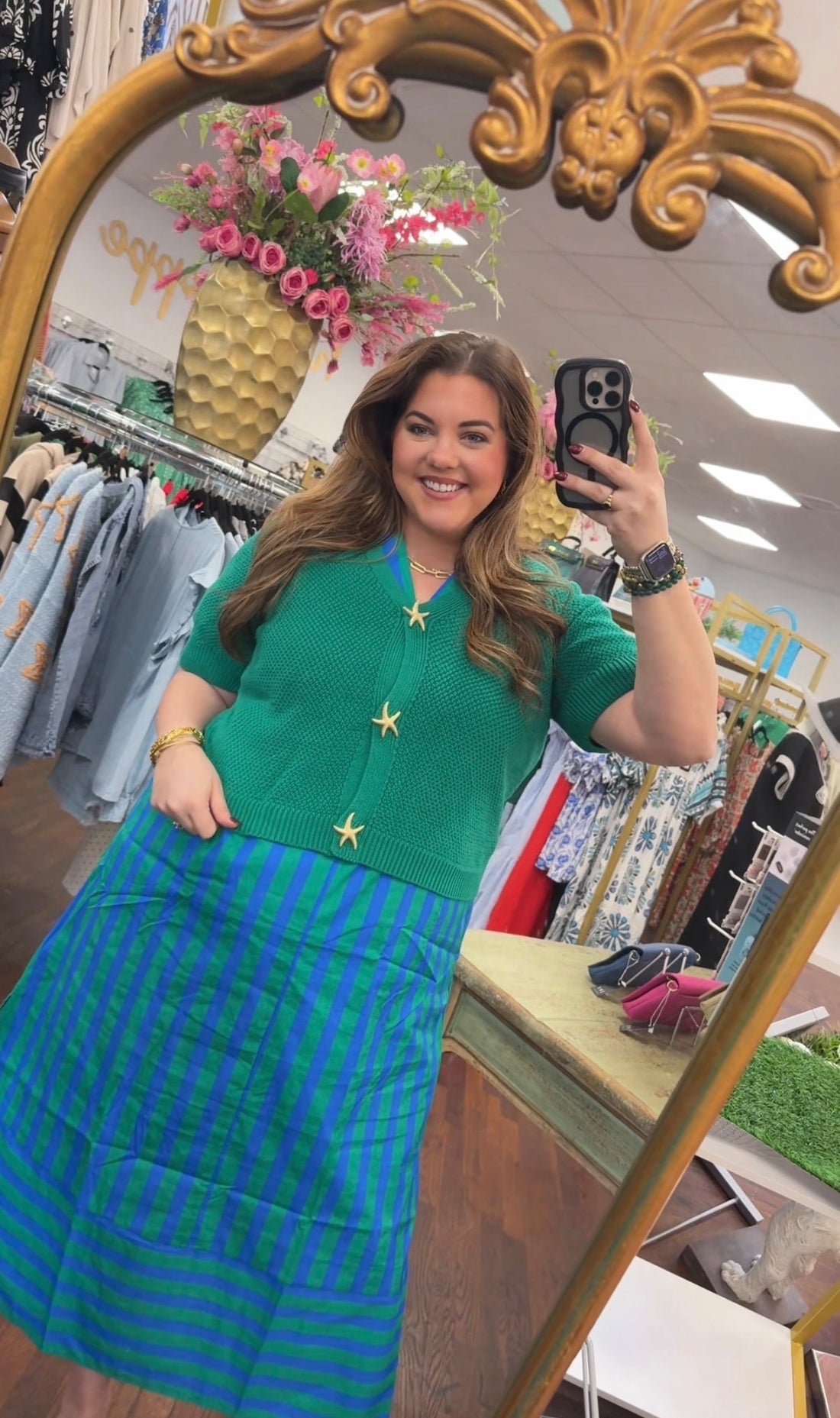 Woman taking a mirror selfie in a clothing store wearing a green top and blue striped skirt.