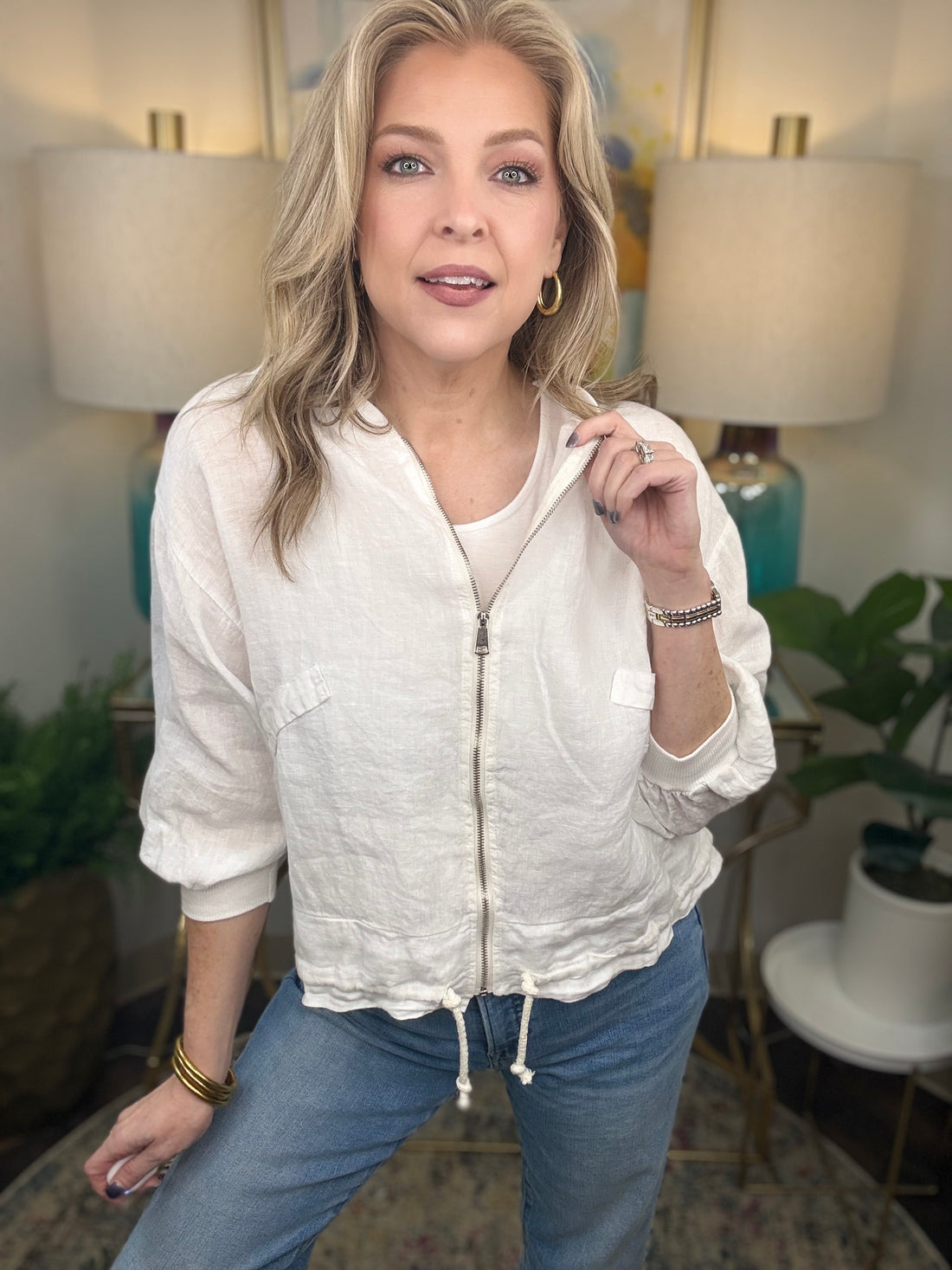 Woman wearing a white zip-up jacket and blue jeans in a room with lamps and plants.