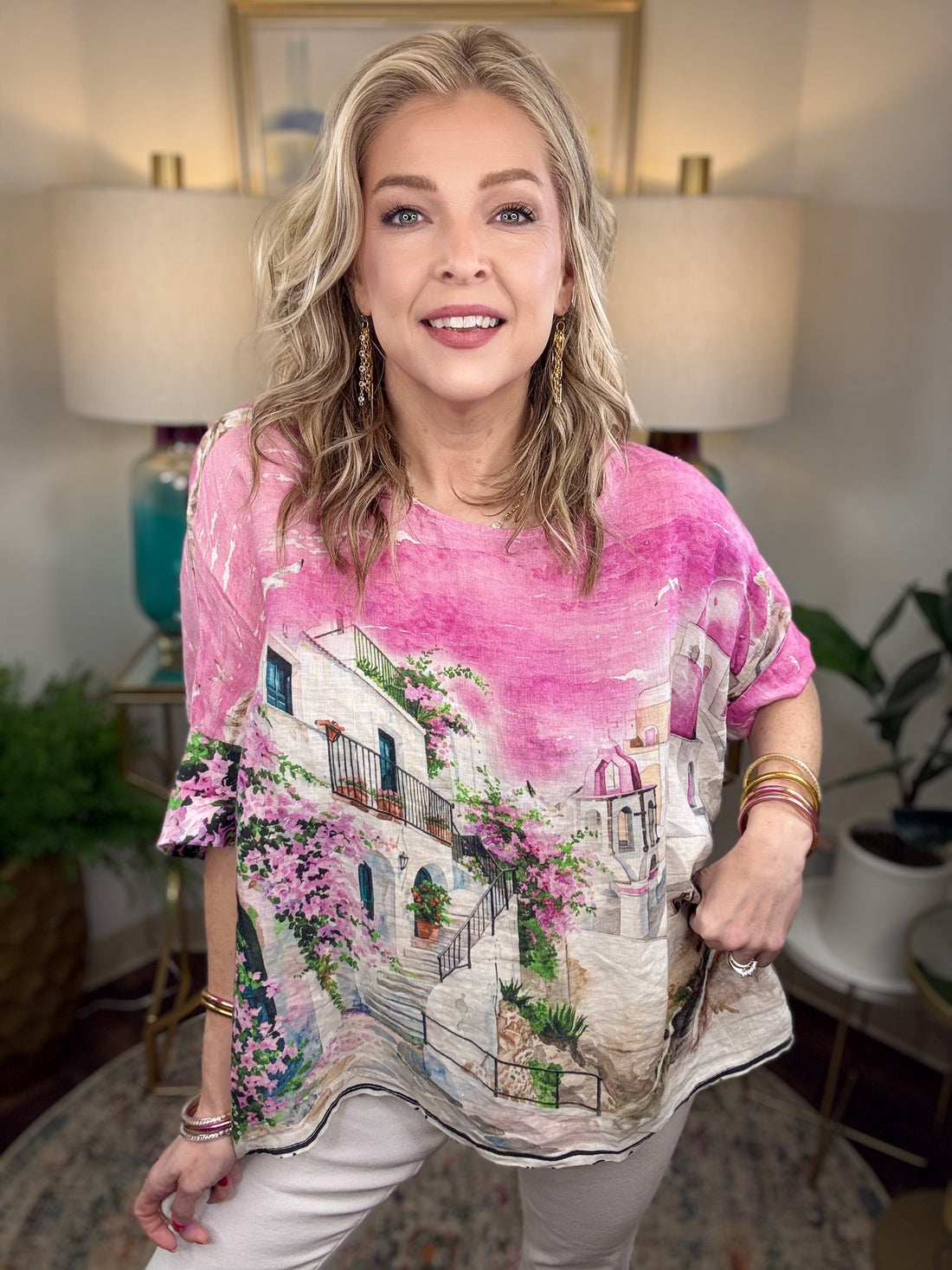 A woman modeling a short-sleeve V-neck top with a pink watercolor wash and a scenic Mediterranean-style village print featuring white buildings, pink flowers, and green vines.