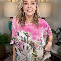 A woman modeling a short-sleeve V-neck top with a pink watercolor wash and a scenic Mediterranean-style village print featuring white buildings, pink flowers, and green vines.