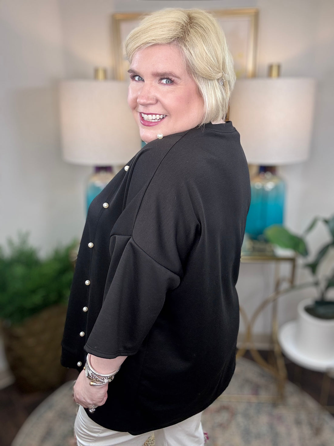 Woman wearing a black tunic top with pearl accents and grommets across the bottom. 