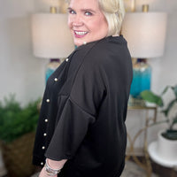 Woman wearing a black tunic top with pearl accents and grommets across the bottom. 