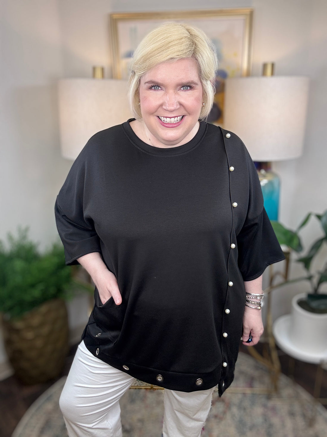 Woman wearing a black tunic top with pearl accents and grommets across the bottom. 