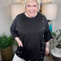 Woman wearing a black tunic top with pearl accents and grommets across the bottom. 