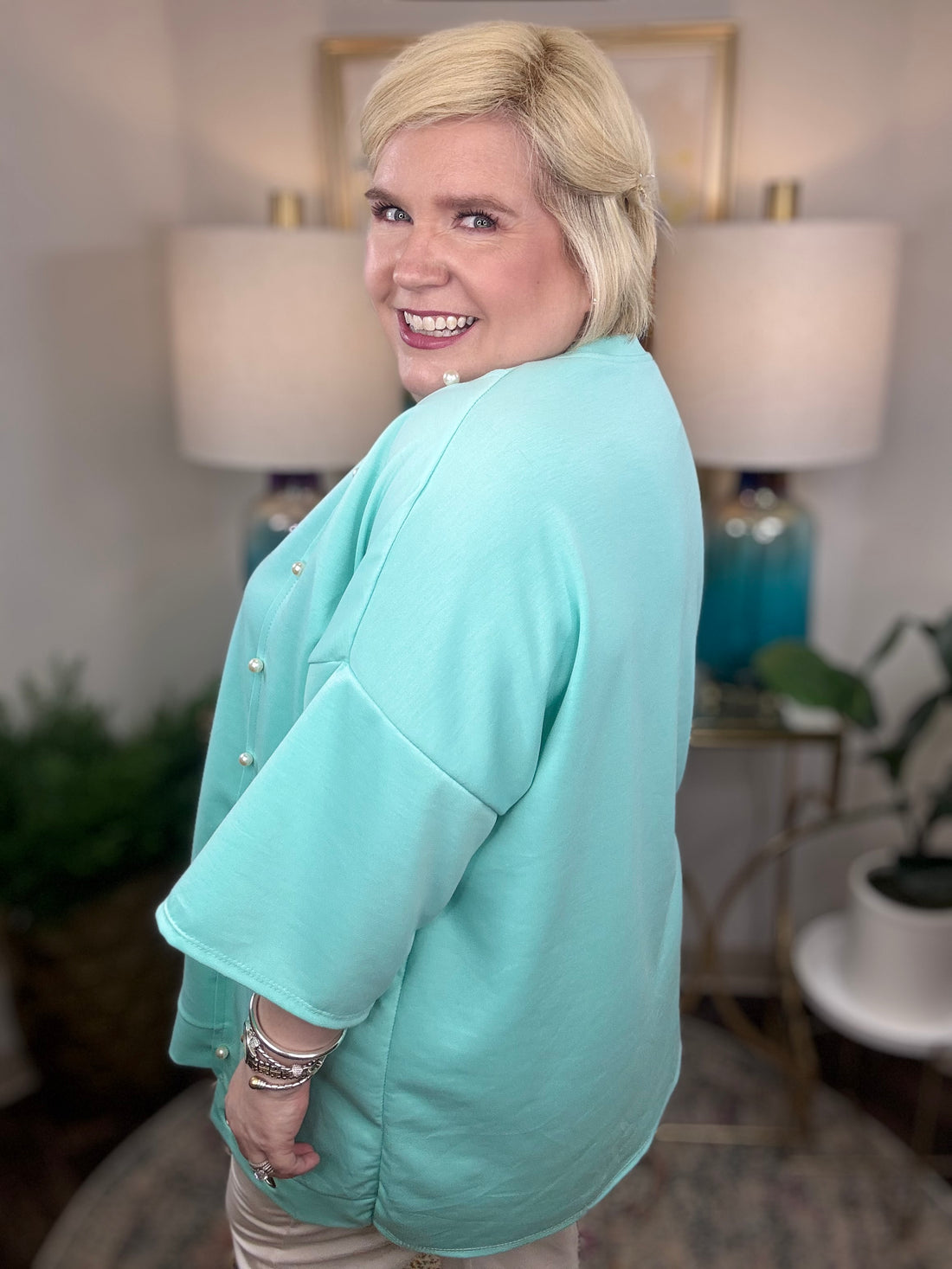 Woman wearing a blue tunic top with pearl accents and grommets across the bottom. 