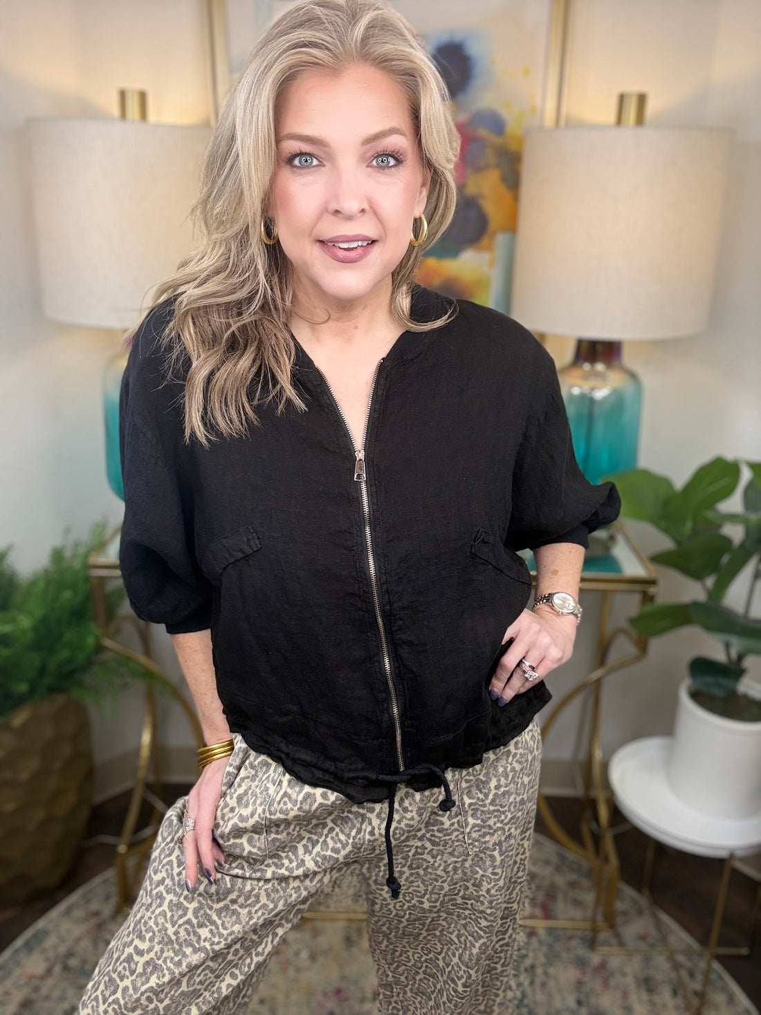 Woman in a black jacket and patterned pants standing in a room with lamps and plants.