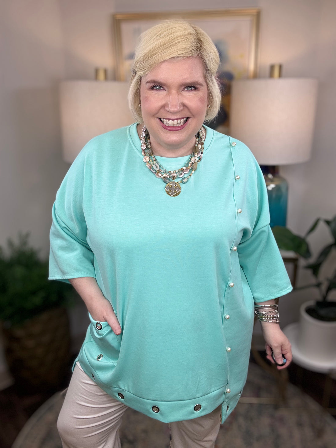 Woman wearing a blue tunic top with pearl accents and grommets across the bottom. 
