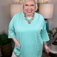 Woman wearing a blue tunic top with pearl accents and grommets across the bottom. 