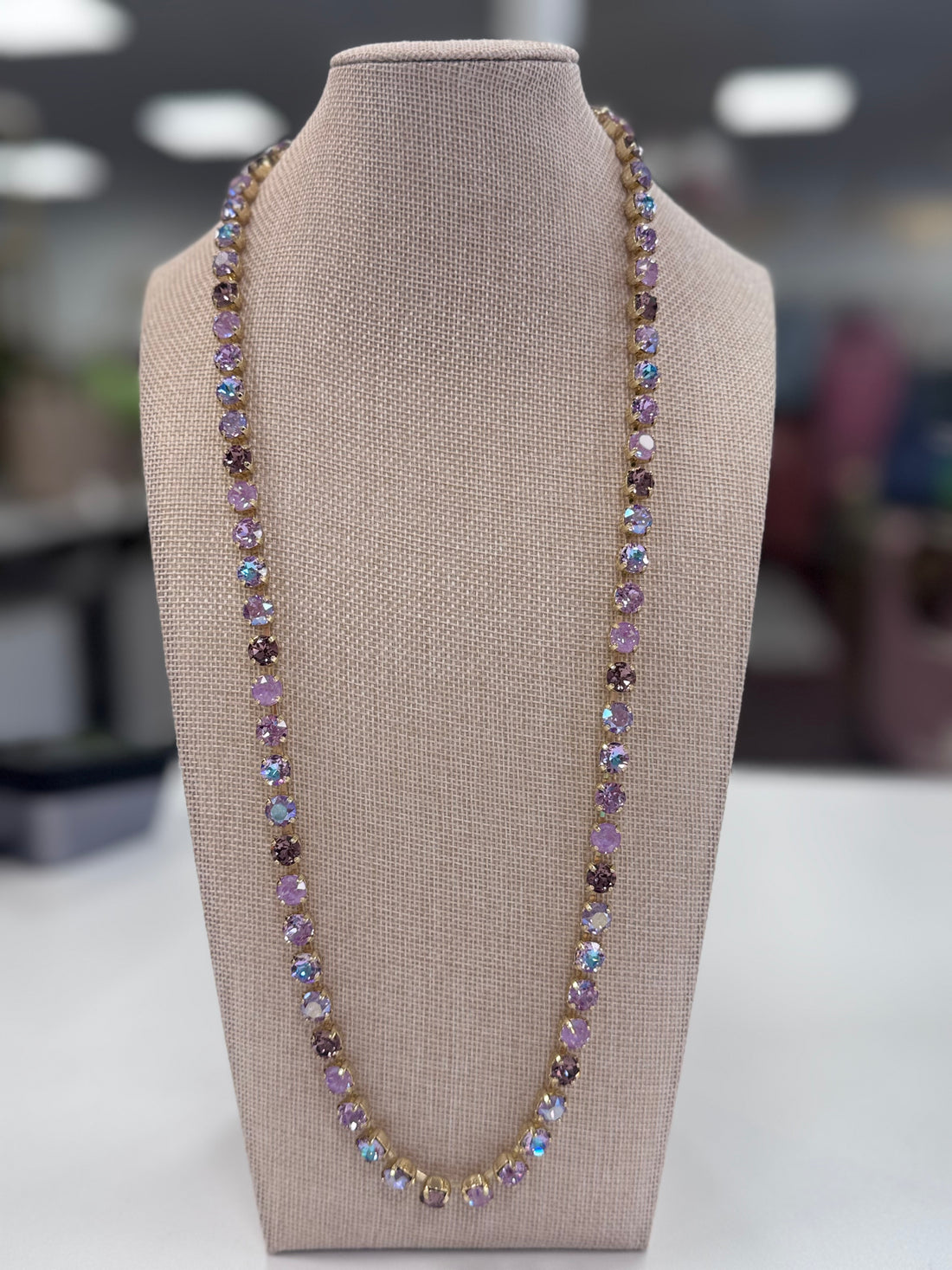 A purple necklace with iridescent stones displayed on a mannequin bust, set against a blurred indoor background.