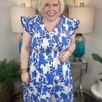 A woman with blonde hair wearing a white and blue floral print midi dress with smocked bodice and ruffled cap sleeve