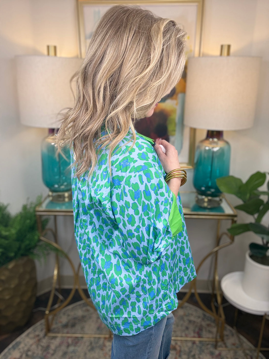 Woman wearing a green and blue patterned shirt in a home setting with lamps and artwork.
