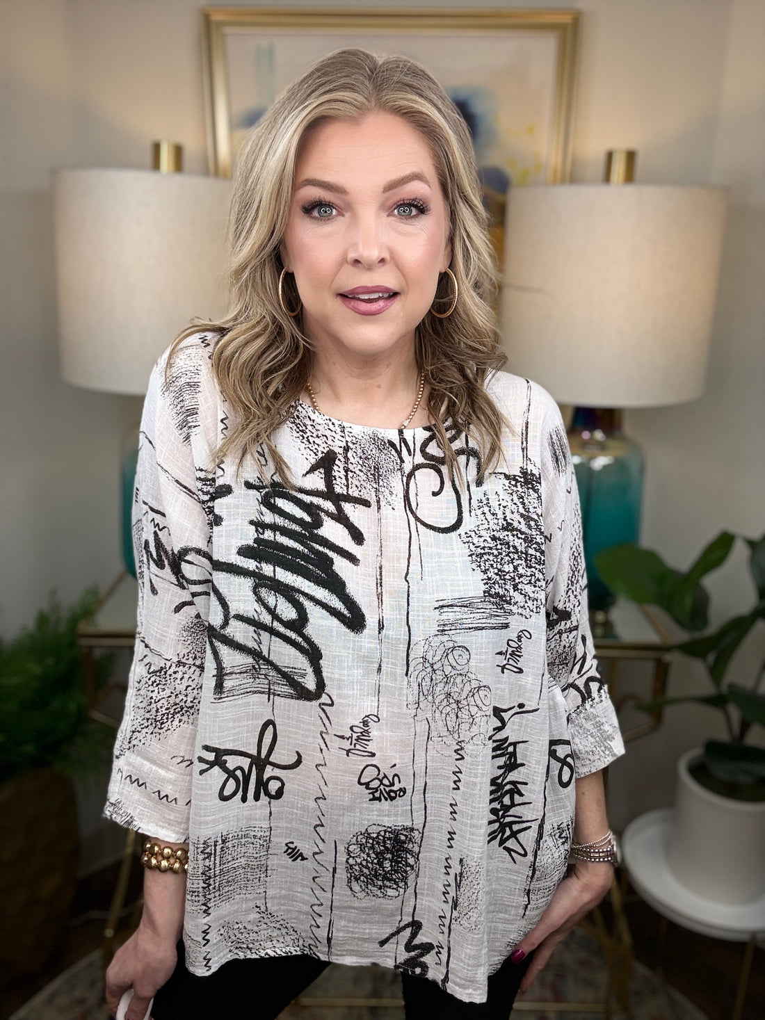 Woman wearing a white blouse with black text and graphics in a room with lamps and decor.