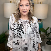Woman wearing a white blouse with black text and graphics in a room with lamps and decor.