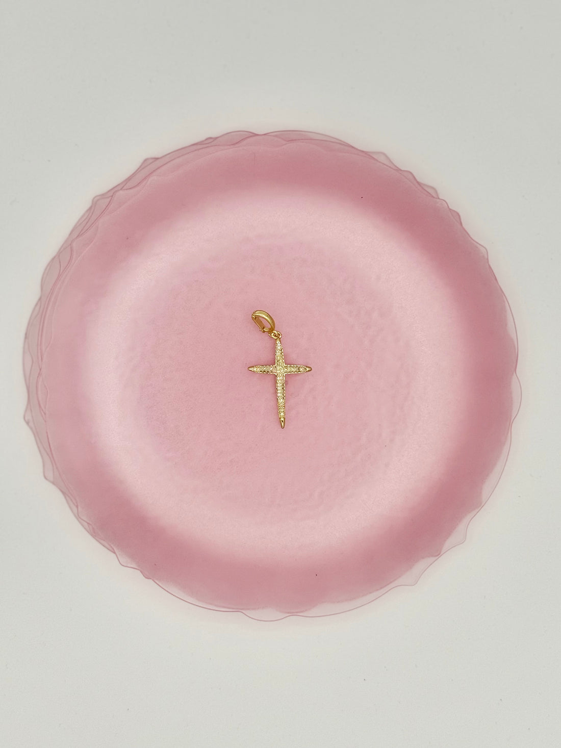A small gold cross pendant with a textured, sparkling surface, displayed on a scalloped pink plate against a light gray background.