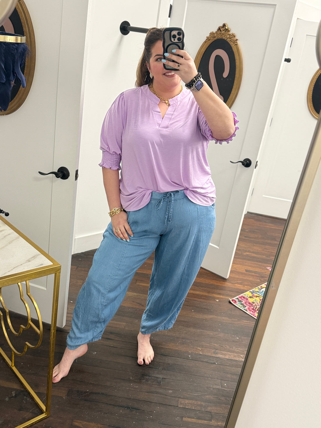Woman taking a mirror selfie wearing a light purple top and blue jeans in a room with a white door and wooden floor.