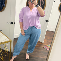 Woman taking a mirror selfie wearing a light purple top and blue jeans in a room with a white door and wooden floor.
