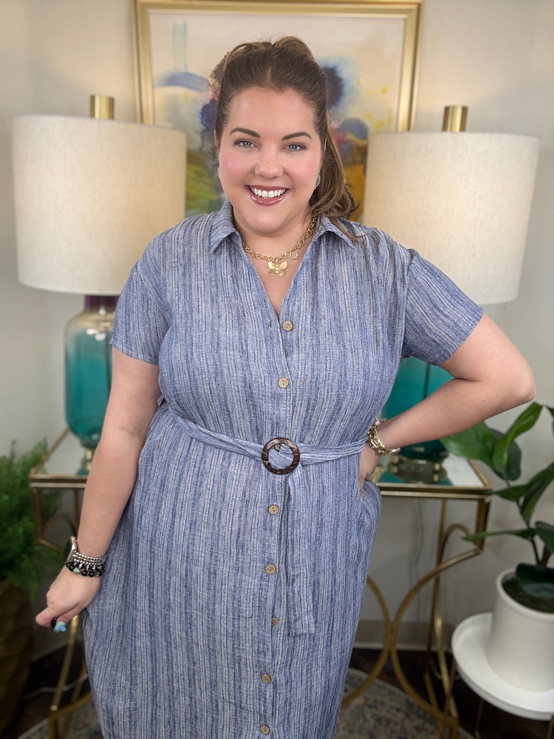Woman wearing a blue dress with a belt, standing in a room with lamps and artwork.