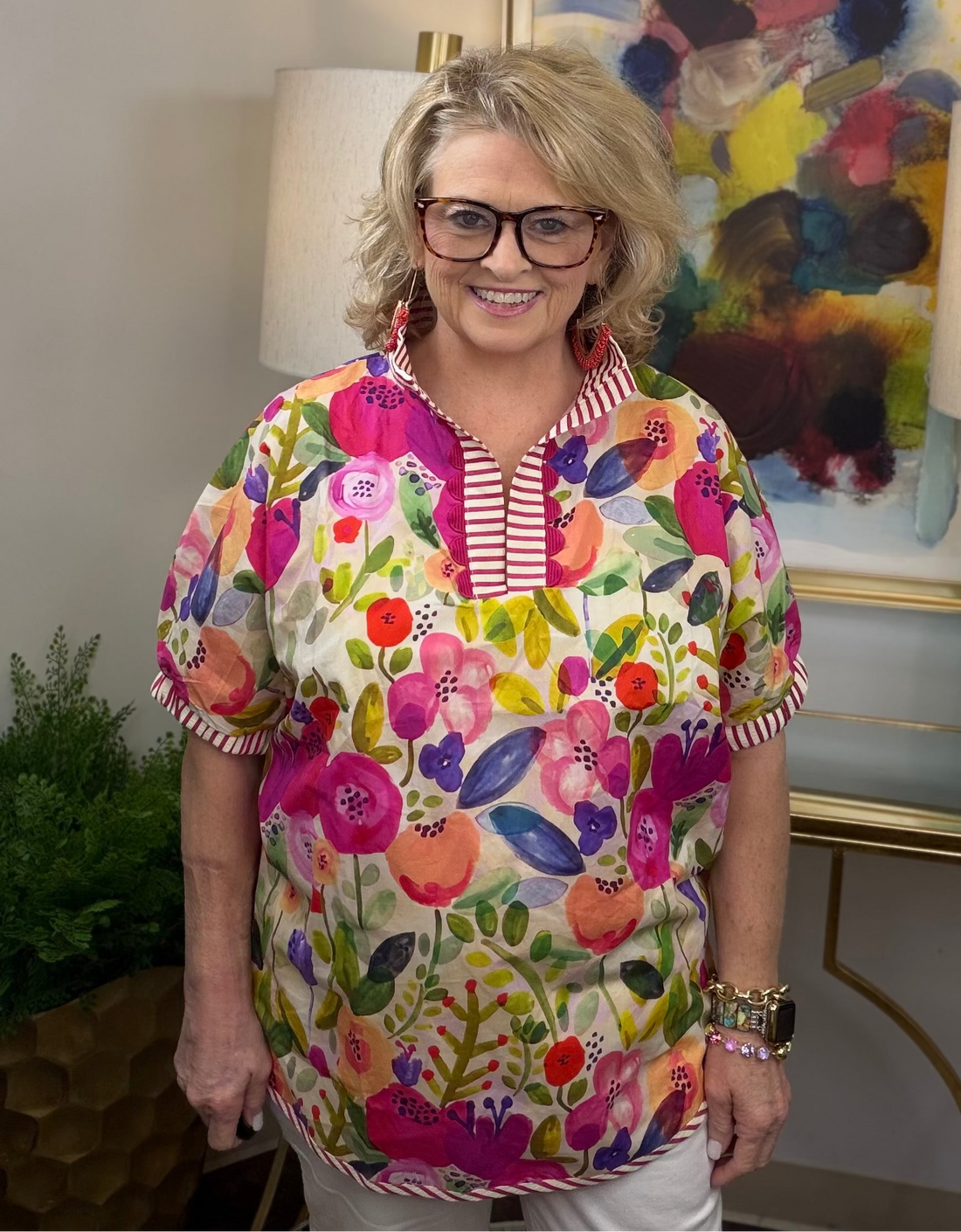 Woman wearing a colorful floral blouse in an indoor setting with a lamp and painting in the background.
