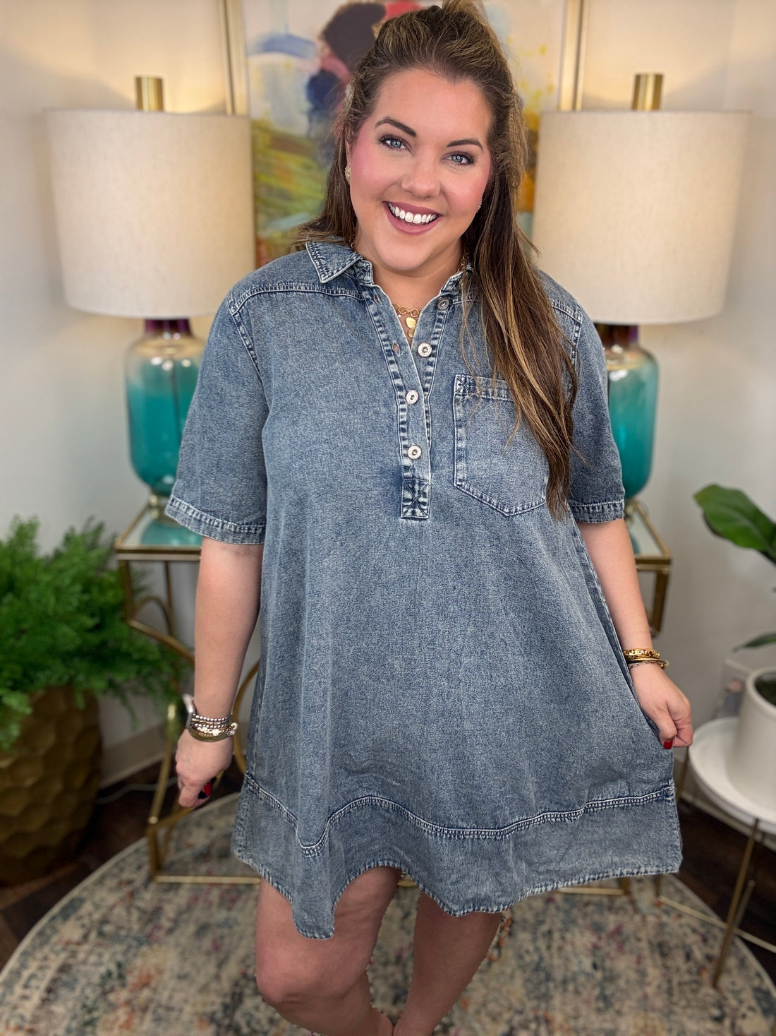 Woman wearing a denim dress in a home setting with lamps and decor.