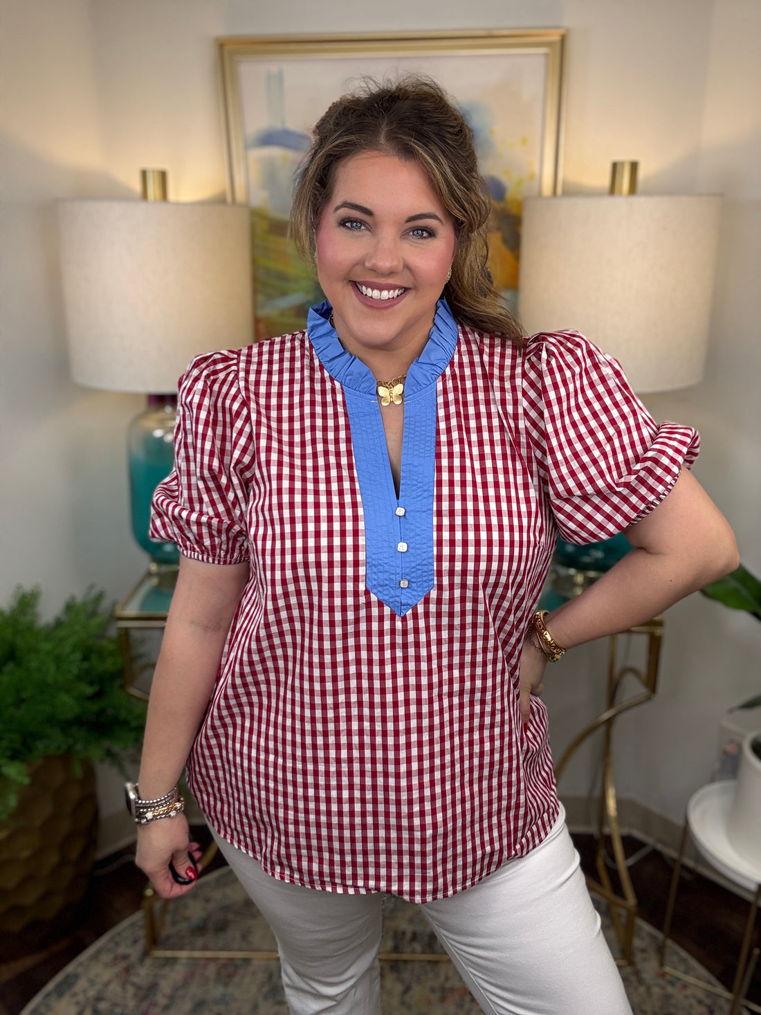 Woman wearing a red and white checkered blouse with blue trim in a room with lamps and a painting.
