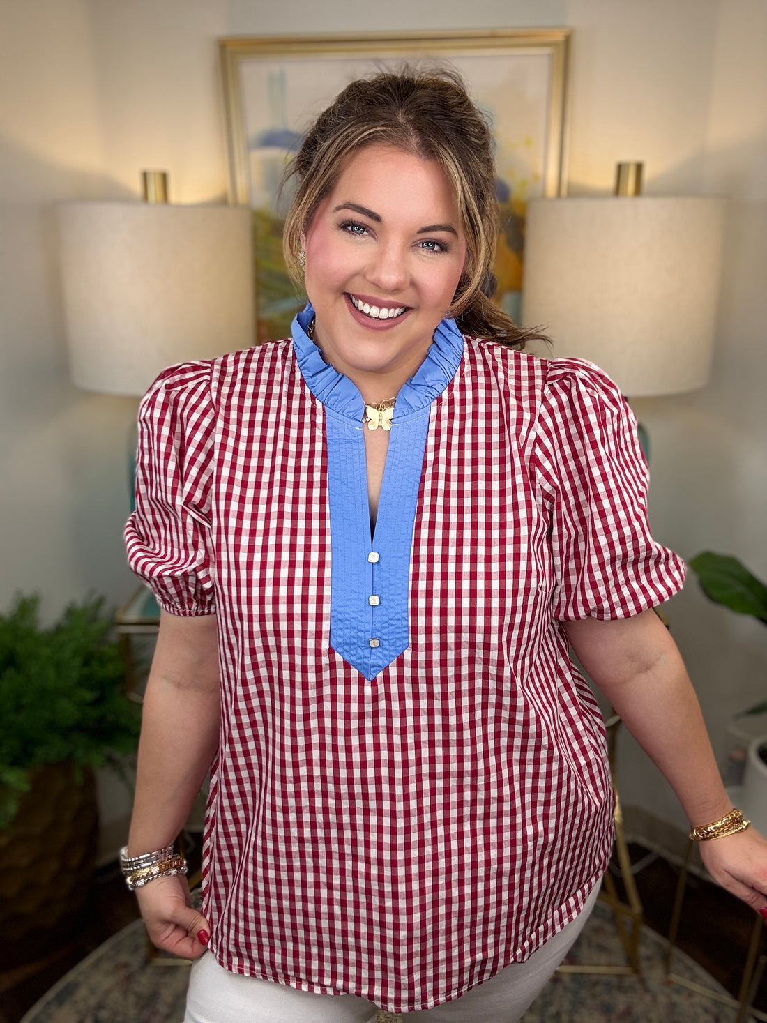 Woman wearing a red and white checkered shirt with blue trim in a room with lamps and a painting.