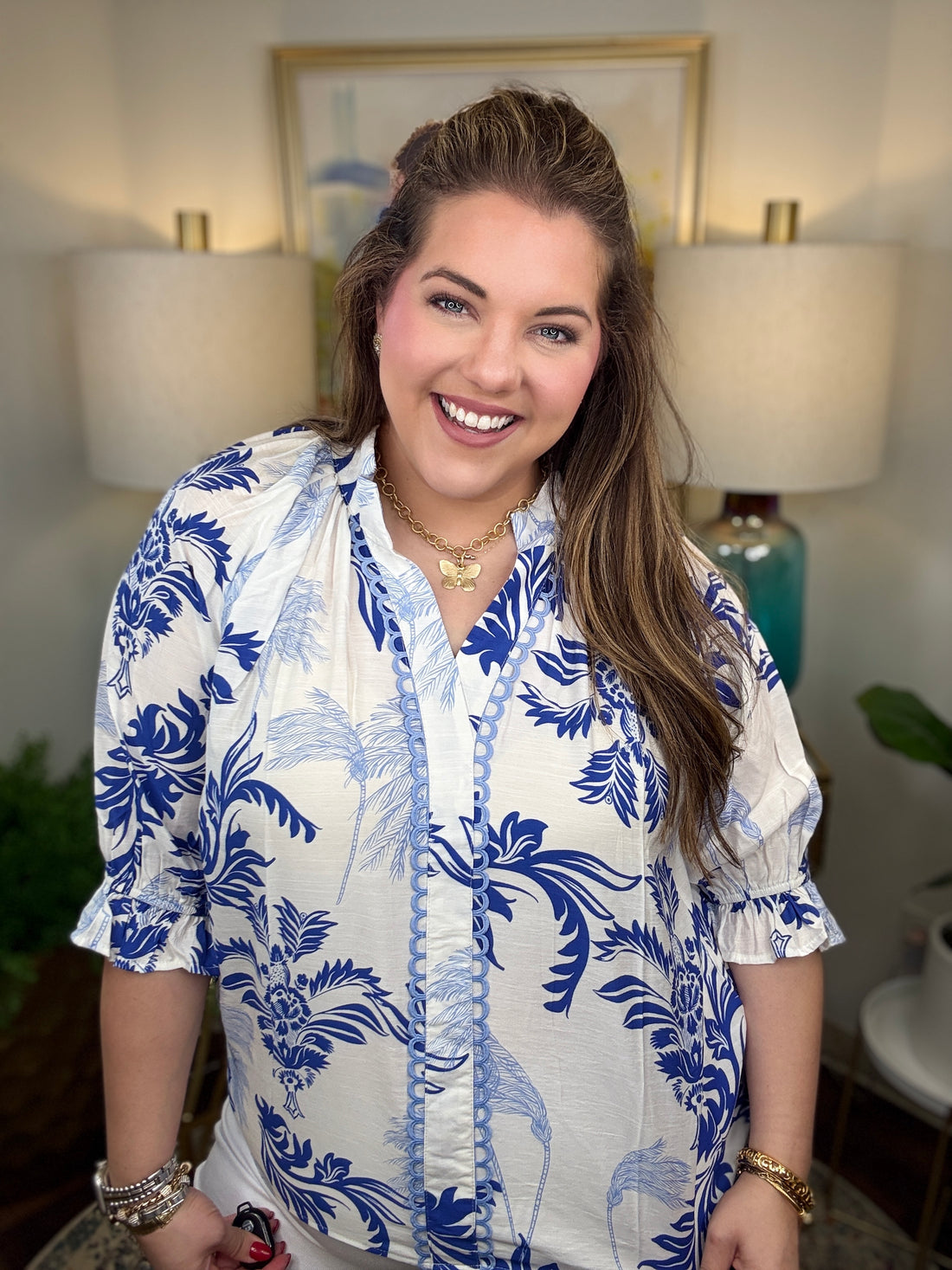 Woman wearing a white blouse with blue floral patterns in a room with lamps and a painting.