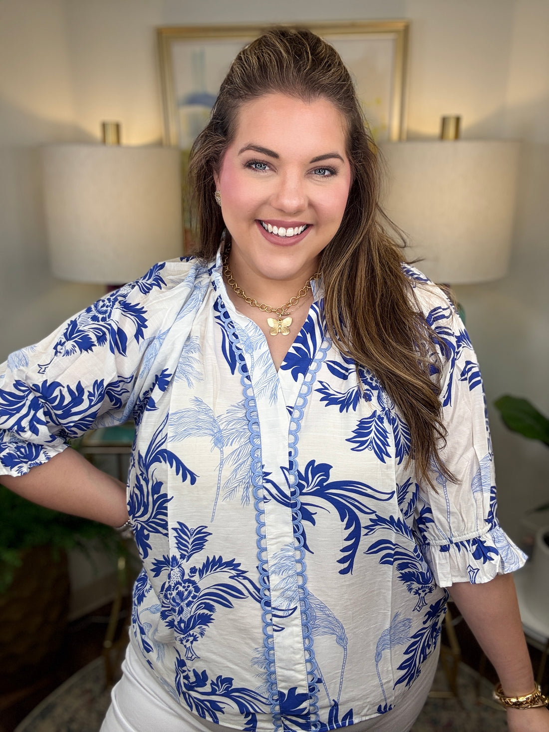 Woman wearing a blue and white patterned blouse indoors with lamps and a painting in the background.