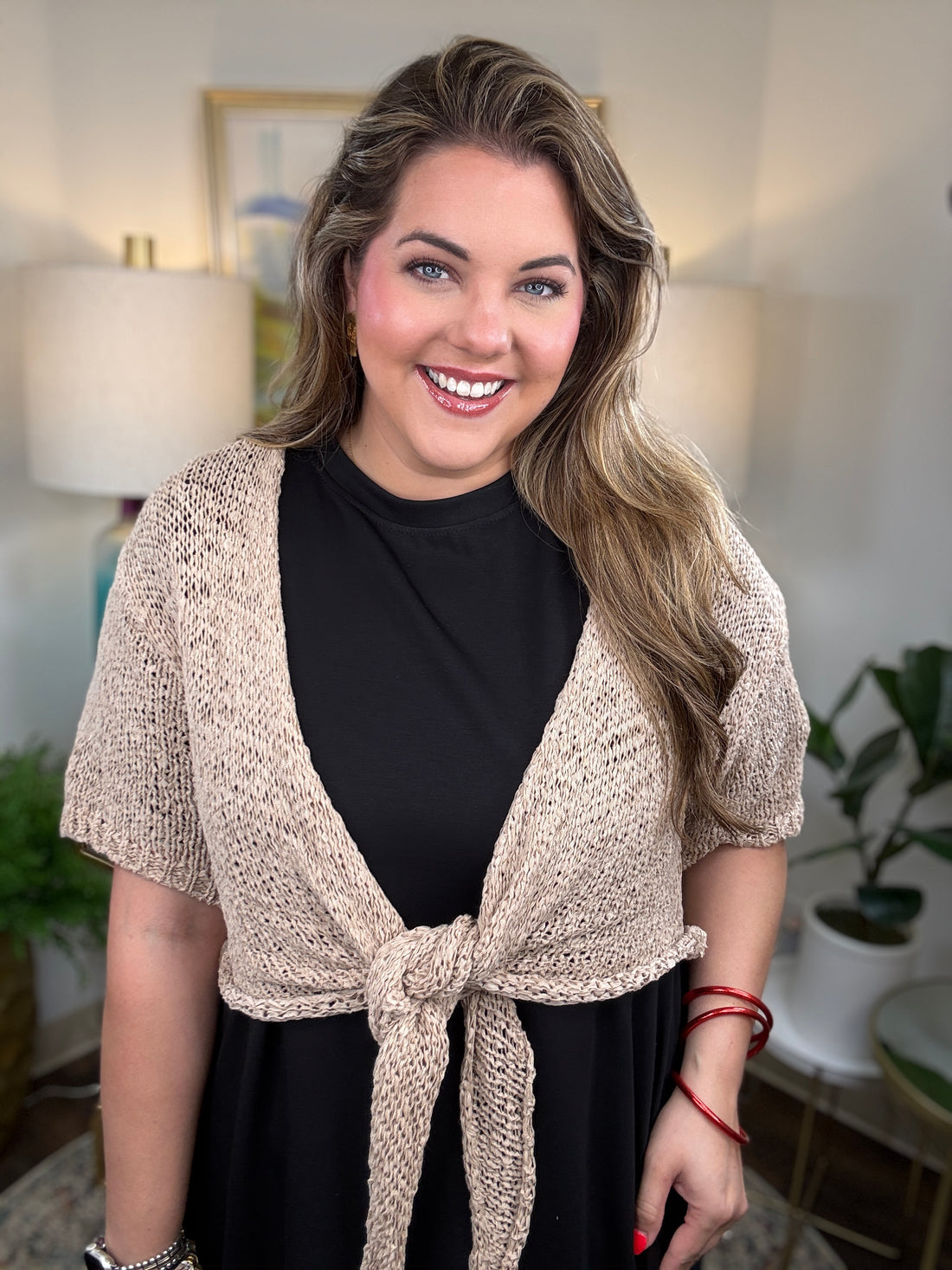 A woman modeling a short-sleeve knit wrap in a warm beige/cream color tied at the waist over a black dress