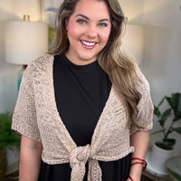 A woman modeling a short-sleeve knit wrap in a warm beige/cream color tied at the waist over a black dress