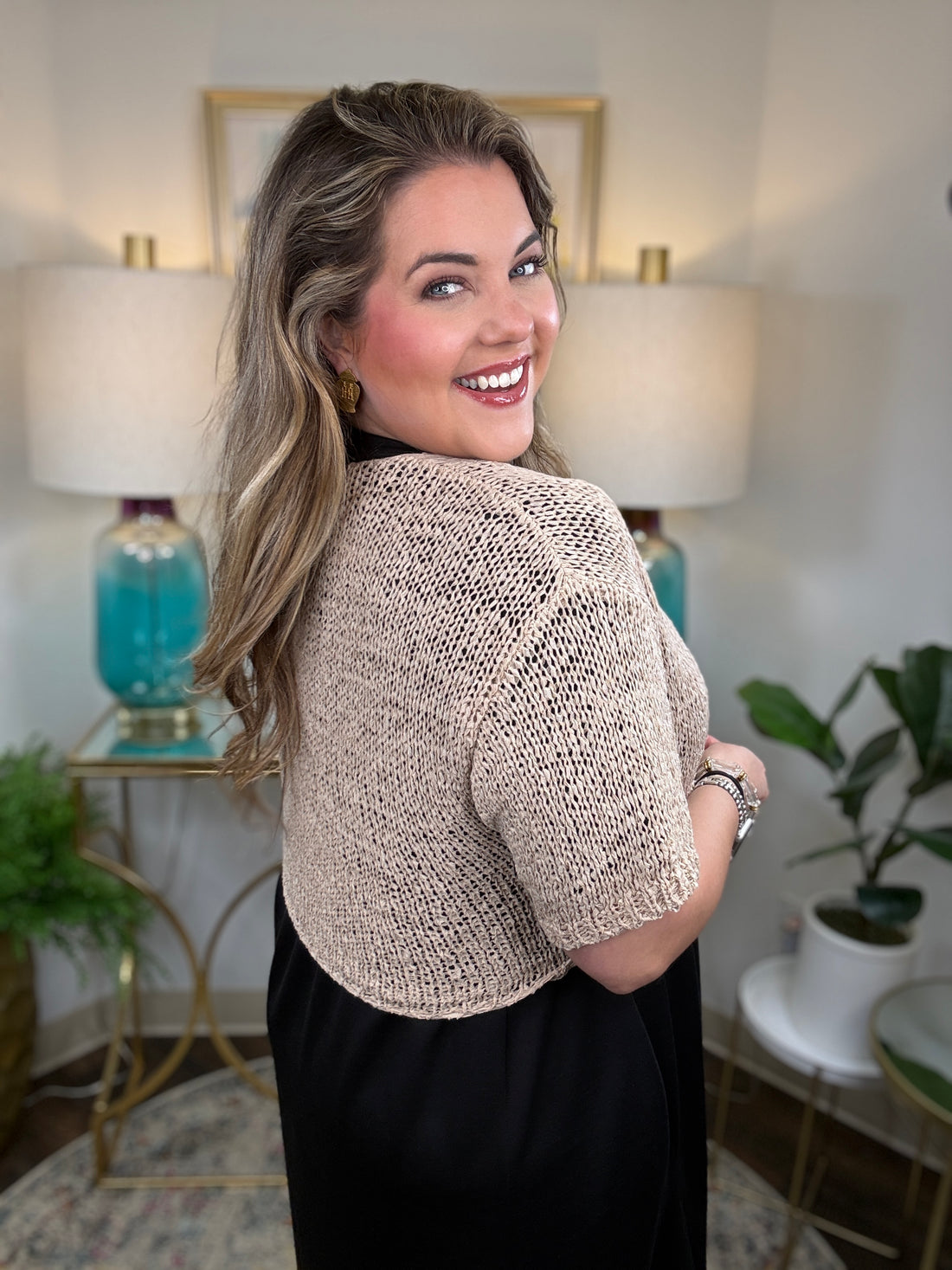 A woman modeling a short-sleeve knit wrap in a warm beige/cream color tied at the waist over a black dress