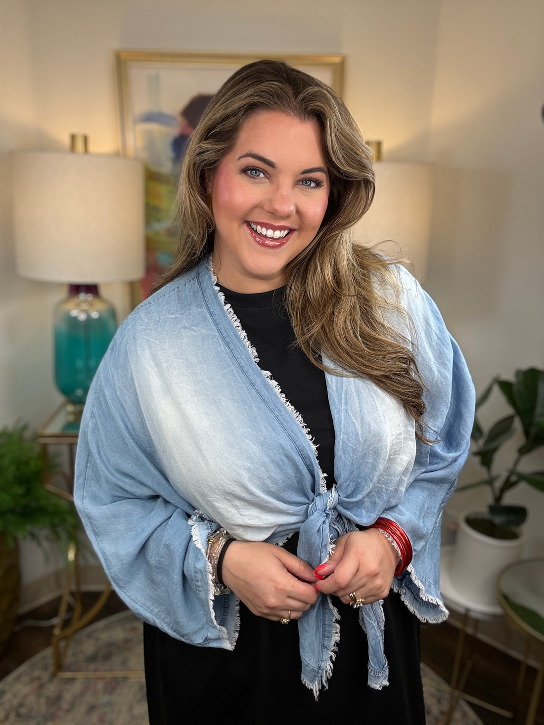 A woman smiling and wearing a denim shrug tied at the waist over a black dress.