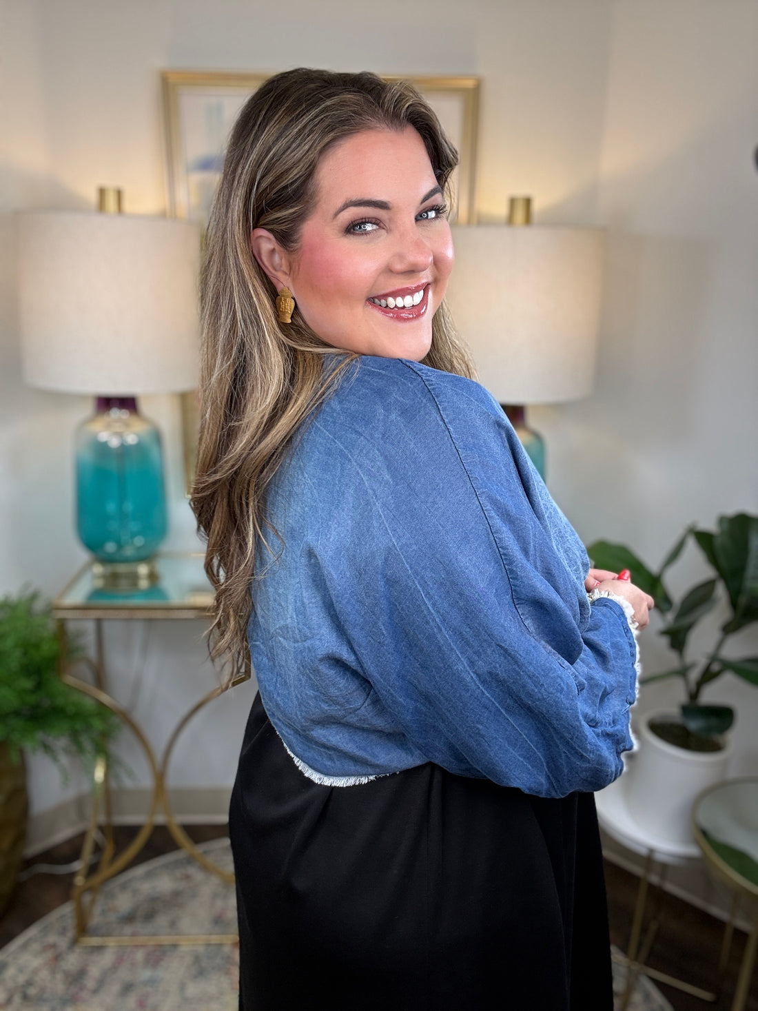 A woman smiling and wearing a denim shrug tied at the waist over a black dress.