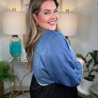 A woman smiling and wearing a denim shrug tied at the waist over a black dress.