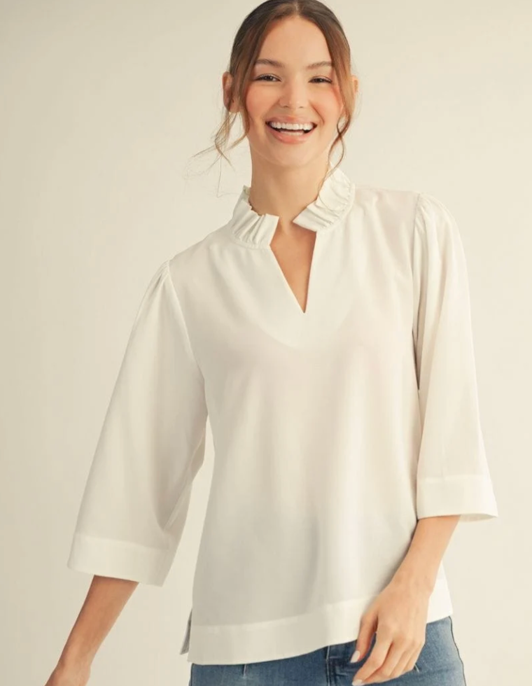 A woman smiling while wearing a cream-colored blouse with a ruffled mock neck and elbow-length sleeves, paired with blue jeans, against a plain light beige background.